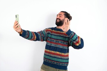 Caucasian man with beard wearing sweater over white background holds modern mobile phone and makes video call waves palm in hello gesture. People modern technology concept