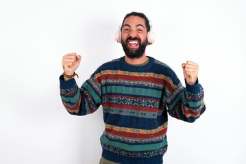 Naklejka premium Emotional Caucasian man with beard wearing sweater exclaims loudly feels like winner raises clenched fists keeps mouth opened wears stereo headphones on ears makes yes gesture, listens favorite music