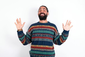 Delighted positive Caucasian man with beard wearing sweater over white background opens mouth  and arms palms up after having great result