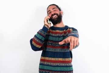 Positive Caucasian man with beard wearing sweater over white background indicates directly at camera has telephone conversation smiles broadly enjoys talking long hours. You join me