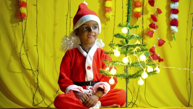 Asian Little Boy In Red Christmas Costume With Gift On Yellow Colored Background. Portrait Of A Satisfied Little Child Boy In Christmas Santa Hat. Laughing Isolated Over Yellow Background.