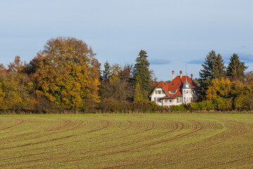 Old villa in autumn. Hidden behind the newly sown field between brightly colored trees is an old...