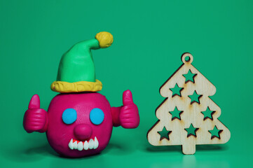 A virus figurine in a festive hat with thumbs up. Next to the Christmas tree. Green background. Christmas toys.
