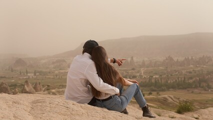 Couple of man and woman sit and hug on mountain slope above valley