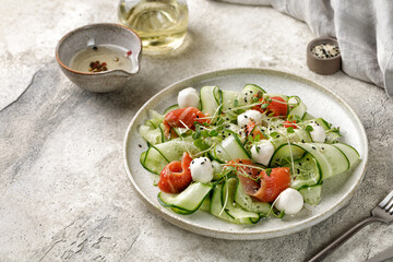 Salmon salad with sliced cucumbers, mozzarella cheese, microgreens on grey ceramic plate and stone background. Diet food concept.