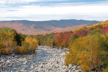 Obraz premium Scenic view of distant mountains in White Mountain National Forest and vibrant fall foliage along banks of rocky Pemigewasset River in New Hampshire.