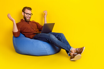 Full length photo of positive man sit comfy bag speak video call netbook isolated on yellow color background