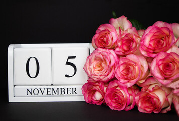November 5 wooden calendar, white on a black background, pink roses lie nearby. Postcard with copy space. The concept of a holiday, congratulation, invitation, party, announcement, vacation,promotion.