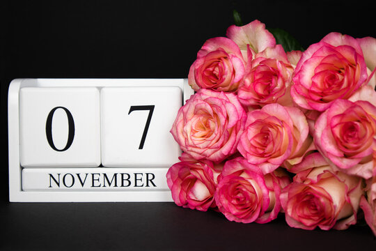November 7 wooden calendar, white on a black background, pink roses lie nearby. Postcard with copy space. The concept of a holiday, congratulation, invitation, party, announcement, vacation,promotion.