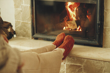 Heating house in winter with wood burning stove. Woman in cozy wool socks warming up feet at...