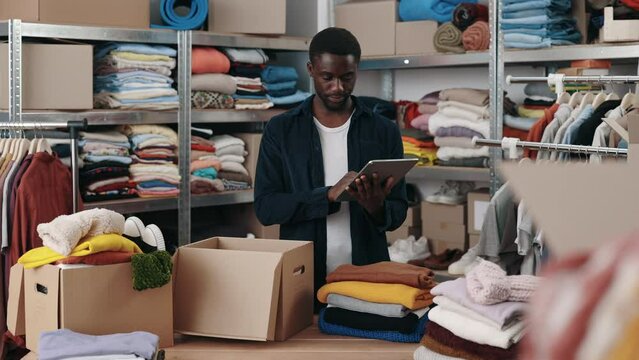 African american male volunteer standing at the warehouse using tablet considering clothes for donations. Belongings at the shelves at the background. Donation and humanitarian aid concept