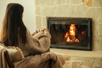 Stylish woman in cozy sweater warming up in chair at fireplace in rustic room. Heating house in...