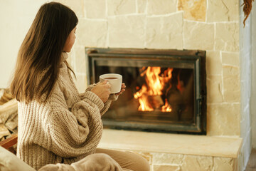 Woman in cozy sweater holding cup of warm tea at fireplace in rustic room. Heating house in winter...