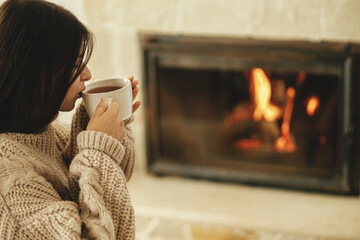 Woman in cozy sweater drinking cup of warm tea at fireplace in rustic room. Heating house in winter...
