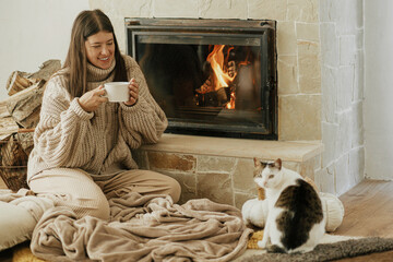 Woman with cup of warm tea and cat relaxing at fireplace, autumn hygge. Heating house with wood...