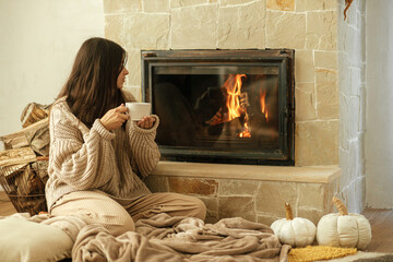 Woman in cozy sweater holding cup of warm tea at fireplace, autumn hygge. Heating house with wood burning stove. Young stylish female relaxing at fireplace in rustic farmhouse