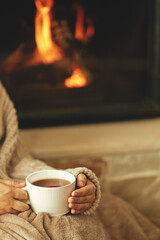 Hands in cozy sweater holding cup of warm tea on background of burning fireplace close up, autumn hygge. Relaxing and warming up at rustic fireplace. Heating house with wood burning stove