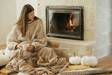 Woman with cup of tea and cute cat sitting together on cozy blanket at fireplace. Adorable tabby...