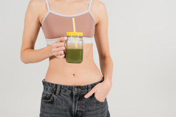 cropped view of young and fit woman holding smoothie while standing with hand in pocket isolated on grey