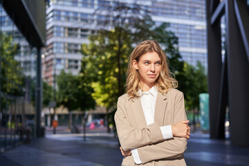 Naklejka na ściany i meble Portrait of young saleswoman, confident businesswoman in suit, cross arms on chest, standing in power pose on street near office buildings