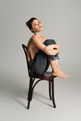 full length of half dressed barefoot woman in jeans sitting on wooden chair on grey background