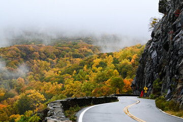 A mountain road in Fall carves into the side of a mountain in front of the edge of the Hudson River...