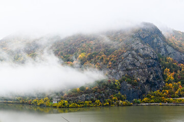 A mountain cliff breaks through the fog overlooking the Hudson River in fall during peak foliage.