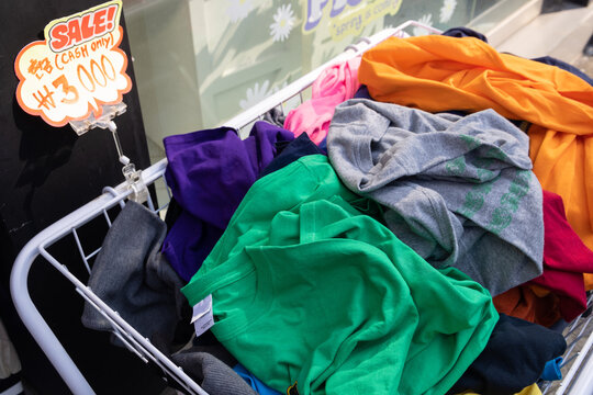 A shopping basket filled with clothes with a note: Sale, cash only. Bunch of t-shirts of different colors for sale. Cheep clothing and fast fashion theme