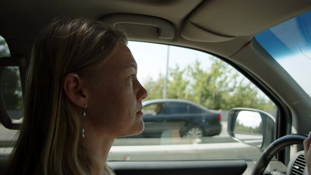 serious young woman driving car along highway ahead and no smile. blonde girl drives rented vehicle on trip and is happy about it. lady holds on steering wheel and looks in rear mirror cheerfully.