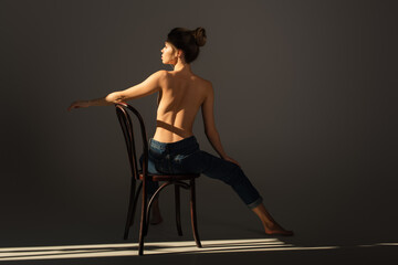 Fototapeta na wymiar back view of half nude barefoot woman in jeans posing on wooden chair on grey background with lighting