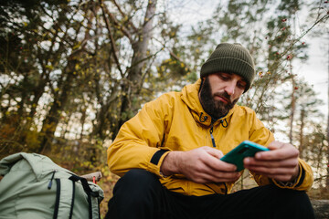 A bearded man rests on a fallen tree in the middle of the forest. A man uses a mobile phone to search GPS.
