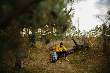 A bearded man rests on a fallen tree in the middle of the forest. A man uses a mobile phone to search GPS.