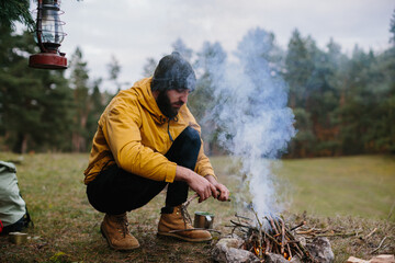 Survival in the wild. A bearded man lights a fire near a makeshift shelter made of pine branches. - obrazy, fototapety, plakaty