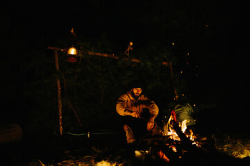 Fototapeta na wymiar The traveler rests by the night campfire. Furnished shelter from branches for sleeping with a kerosene lamp and a fire surrounded by stones.