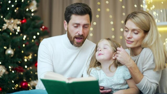 caring parents are reading a book to their little daughter in a cozy house on Christmas Eve, sitting at home on the sofa background New Year tree. Winter holiday evening. Happy family with child