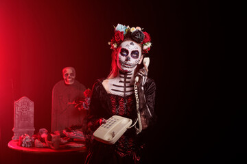 Santa muerte model using landline phone to answer call, holding office cord telephone on mexican traditional holiday. Looking like la cavalera catrina on holiday, celebrating dios de los muertos.