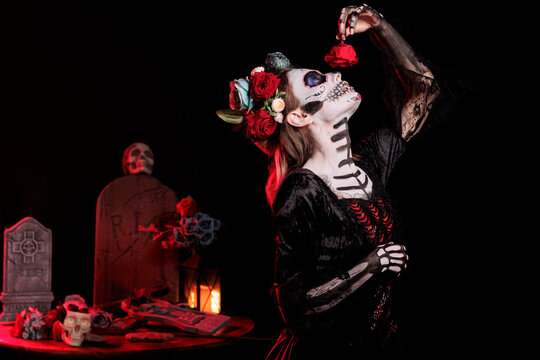Glamour woman smelling roses in studio, posing with flowers and looking like la cavalera catrina on day of the dead. Scary model with costume and skull make up acting like lady of death.