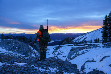 Hunter with rifle dressed in camo stands on the edge of a snowy mountain ridge looking for deer at...