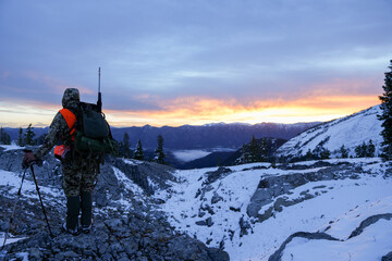 Hunter with rifle dressed in camo stands on the edge of a snowy mountain ridge looking for deer at...