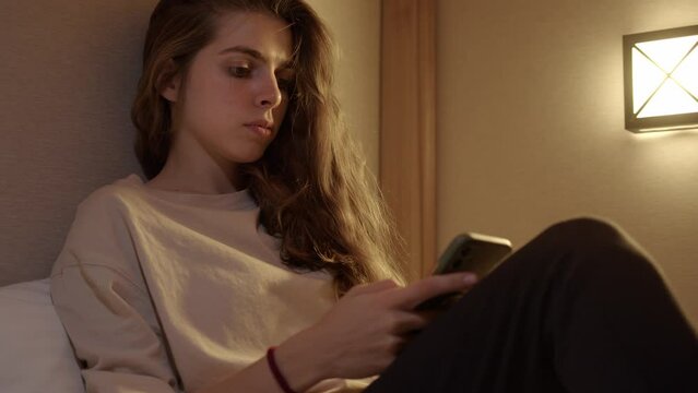 curly young woman lies in bed in evening and surfs Internet on smartphone. girl in white T-shirt scrolls through photos or reads text on phone in hotel room with yellow natural light from light bulb.