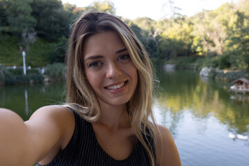 Close up young smiling woman in casual clothes posing with lake. People sincere emotions lifestyle concept. Doing selfie shot on mobile phone.