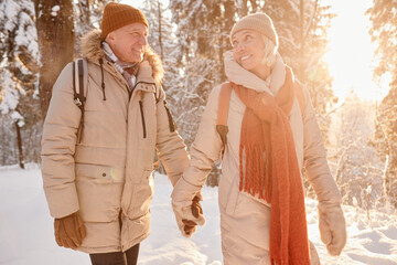 Waist up portrait of happy senior couple enjoying hike in winter forest and holding hands lit by sunlight