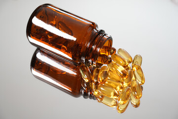 Fototapeta premium Fish oil capsules close-up with selective focus and reflection on a smooth mirror surface. Scattered from brown glass bottle and the half bottle include in screen. Capsules and bottle top-right view.