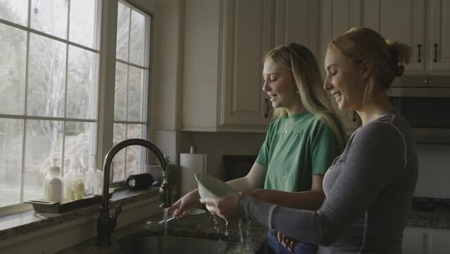 Smiling sisters washing dishes and singing to music in kitchen / Highland, Utah, United States