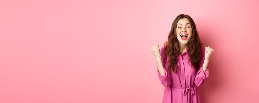 Beautiful smiling woman scream with happy and excited face, saying yes, making fist pump, winning and feeling like champion, triumphing, standing over pink background