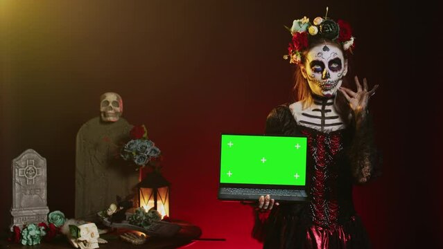 Goddess of death holding laptop with greenscreen, showing isolated copyspace display on pc. Halloween model with creepy body art having isolated chroma key mockup template in studio.