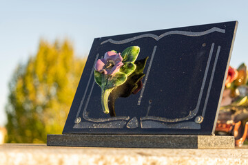 Funeral plaque in the shape of a black book, decorated with a pink flower, blue sky in the background