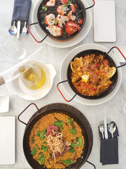 Flatlay of three solo Spanish paella dishes. Paella Negra, Valencia, Chicken on white marble table. Cutlery in table napkin, olive oil in glass bottle.