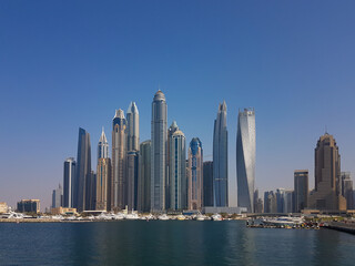 Fototapeta na wymiar Cityscape of tall, modern buildings. Skyscrapers in Dubai Marina on a bright summer day. Clear, blue skies, calm waters, and yachts moored on the docks.