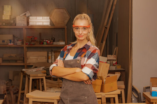 Portrait woman carpenter wearing safety glasses in a furniture manufacturing plant stands in a woodworking workshop holding workshop proudly looking at a smart camera, hands crossed arms.